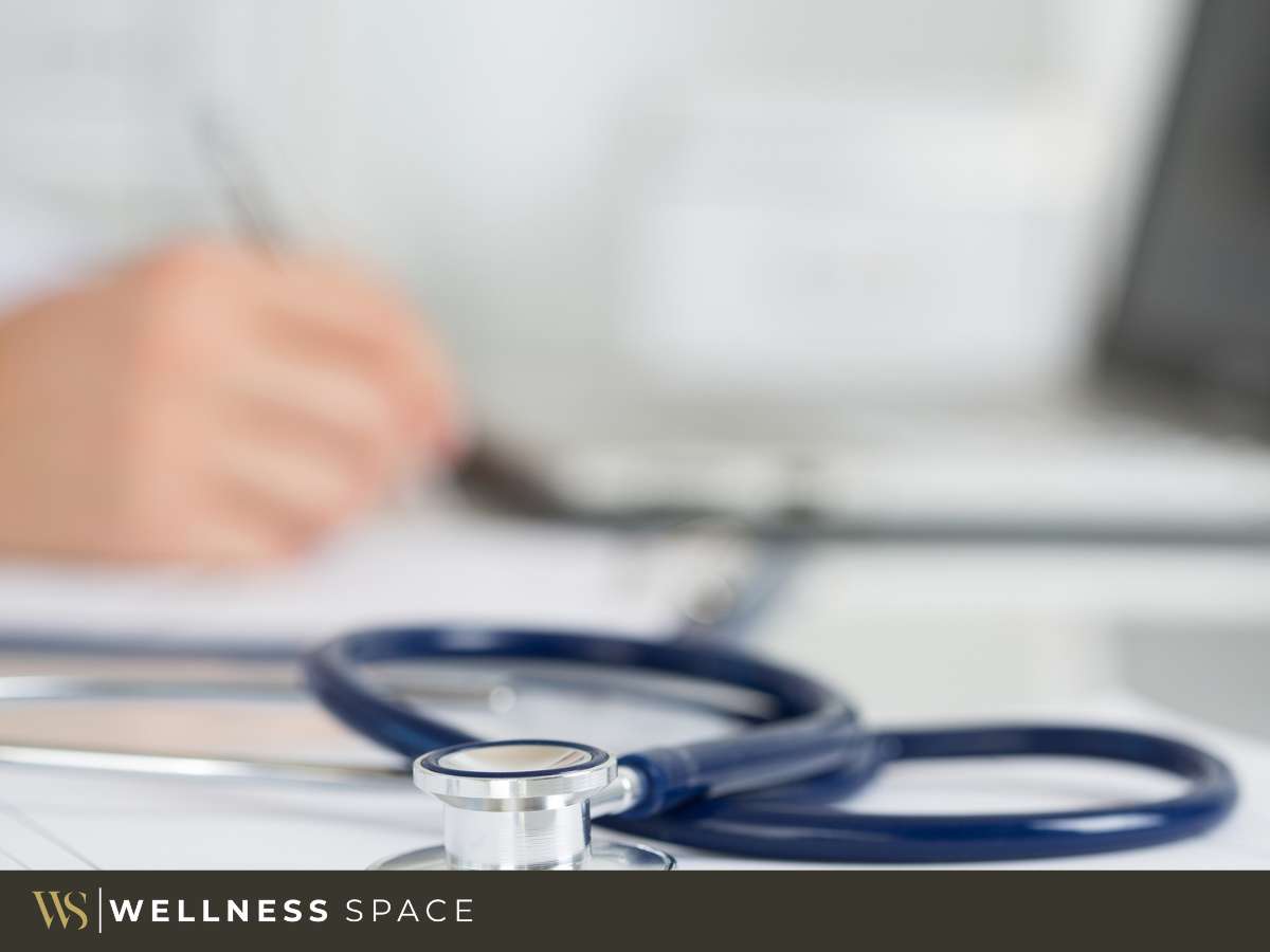 A stethoscope lies on a desk in a medical coworking space, with a blurred doctor writing in the background, symbolizing collaboration in healthcare